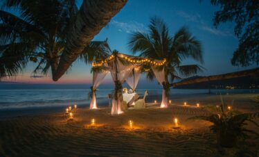 A picturesque romantic dinner setup under a canopy on a Thai beach at twilight, surrounded by candles.